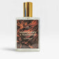 A rectangular glass bottle of perfume with a shiny gold cylindrical cap, featuring a label with a dark brown and orange wood grain pattern and the text 'MAHOGANY & TEAKWOOD EAU DE PARFUM'.