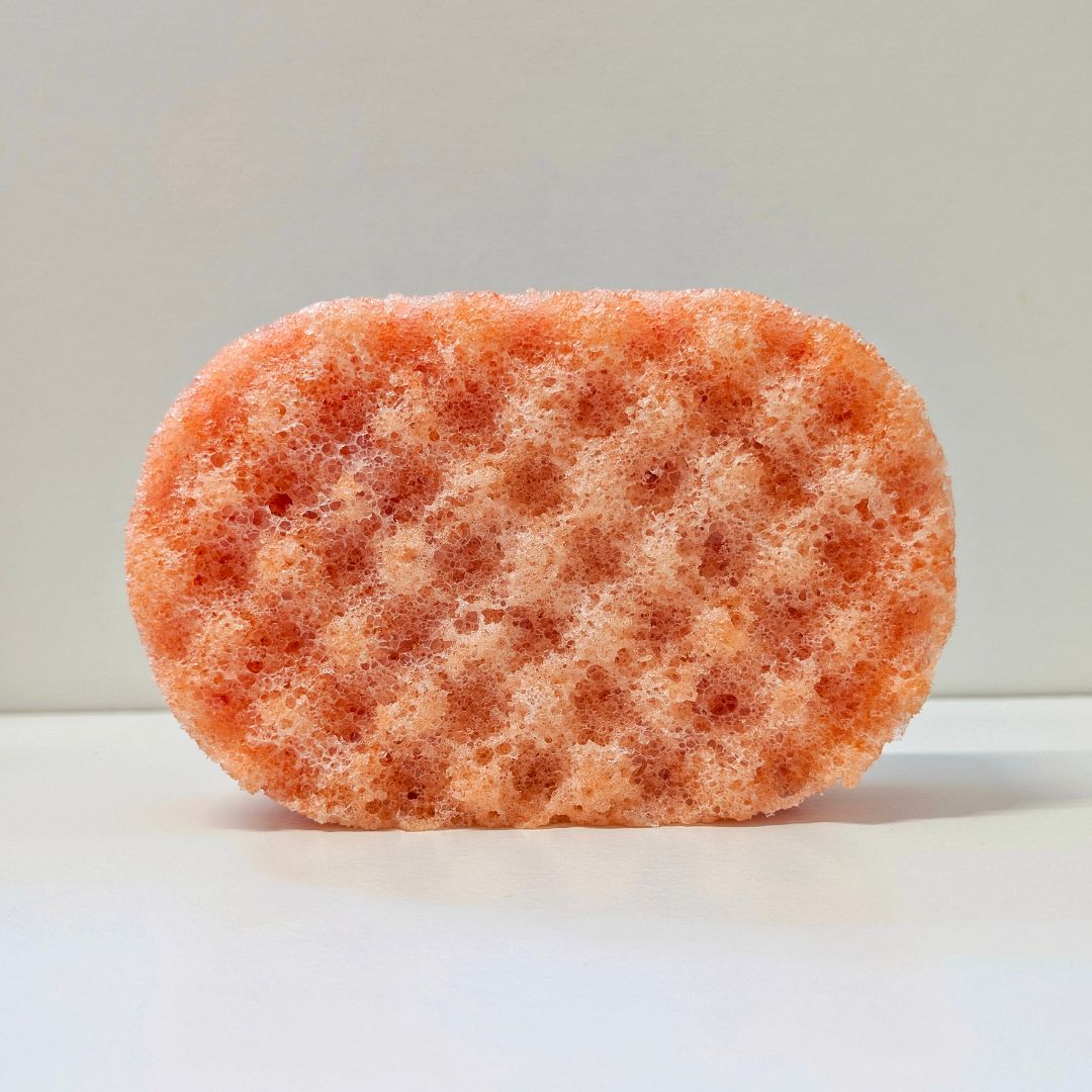 A single oval-shaped orange sponge with a highly porous, textured surface, photographed on a clean white surface against a neutral light gray background.