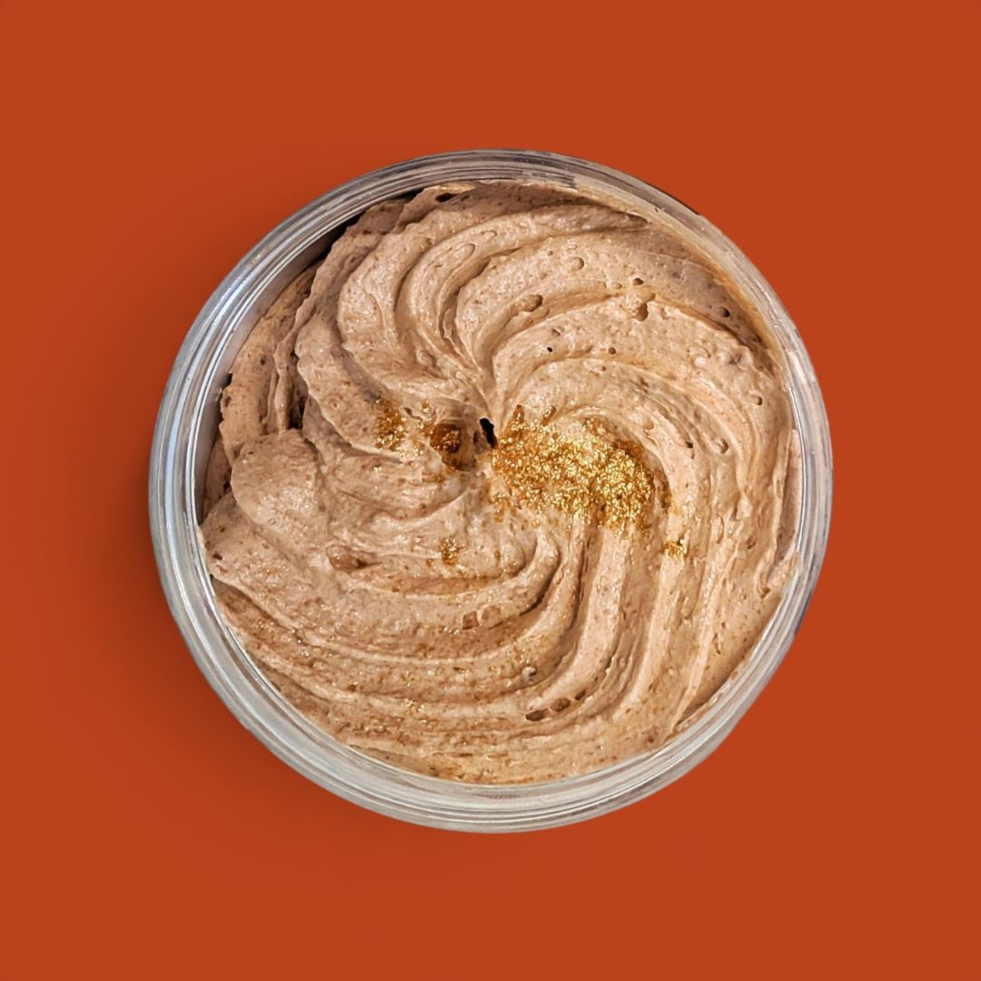 A top-down view of a whipped beige soap swirled inside a clear round container, with a small gold glitter accent on top, set against a solid orange background.