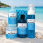 Four Maldives branded self-care products arranged on a sandy beach with turquoise water and seashells in the background: a large blue bottle, a smaller blue jar, a dark bottle with a black cap, and a tall clear bottle with a white pump. A blue sponge is behind them.