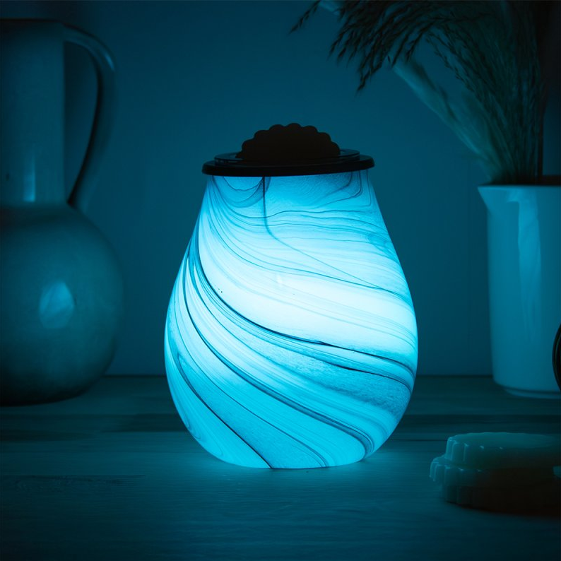 Glowing blue wax melt warmer in a dimly lit room with plants and a plant pot.