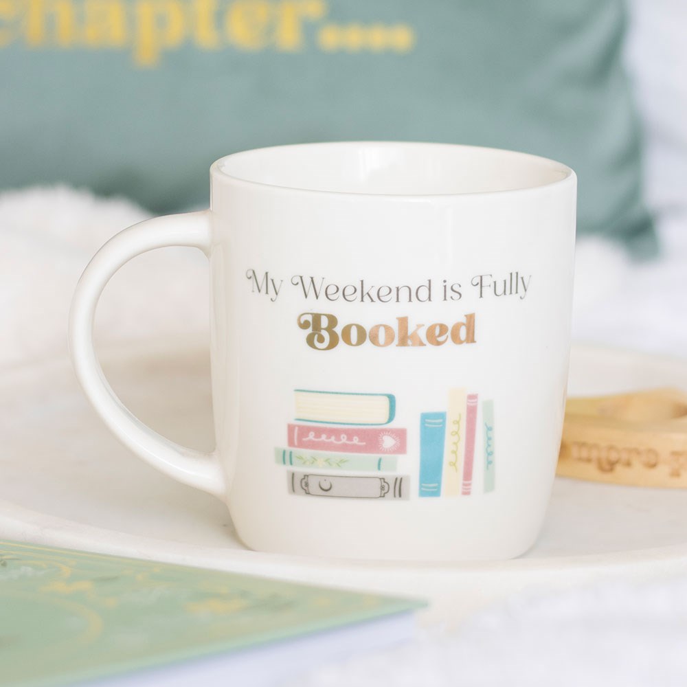 White mug with 'My Weekend is Fully Booked' text and book illustrations on a light surface.