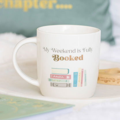 White mug with 'My Weekend is Fully Booked' text and book illustrations on a light surface.