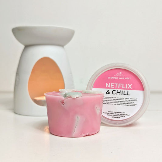 A chunky pink and pale green wax melt next to a pot labelled Netflix & Chill with a white wax melt burner behind. 