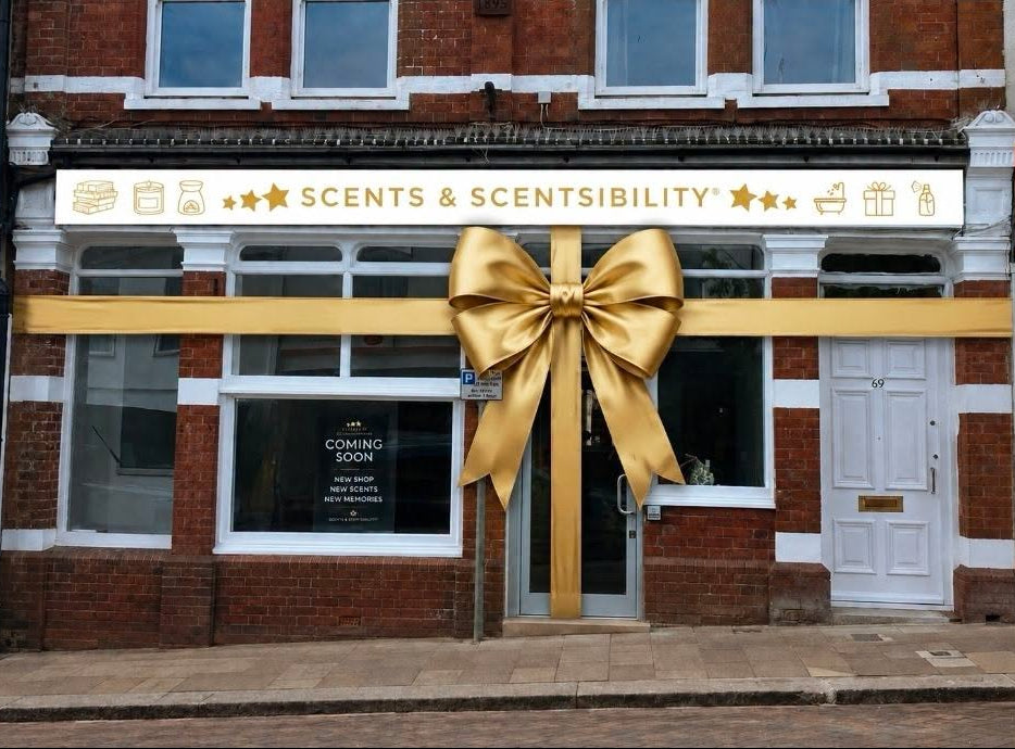 A brick storefront with a large gold ribbon and oversized gold bow across the front door and window area. The shop sign reads 'SCENTS & SCENTSIBILITY' and a window says 'COMING SOON'.