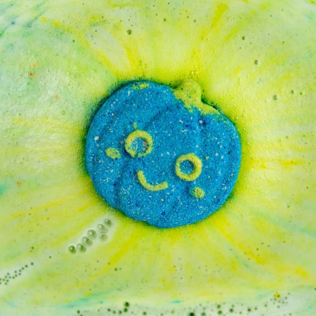 A close-up of a pumpkin shaped blue bath bomb with a yellow smiley face, surrounded by a yellow and green bubbly foam burst.