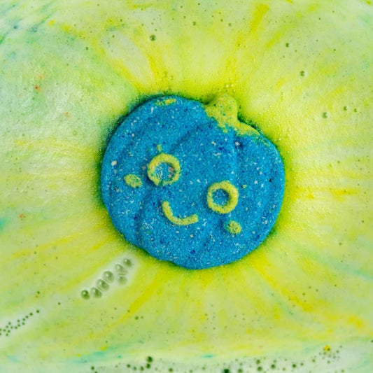 A close-up of a pumpkin shaped blue bath bomb with a yellow smiley face, surrounded by a yellow and green bubbly foam burst.