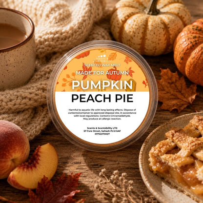 A round plastic tub of Scents & Scentsibility 'Pumpkin Peach Pie' scented wax melt sits on a rustic wooden table surrounded by autumn-themed props including a mug of coffee, peaches, a pie, and pumpkins.