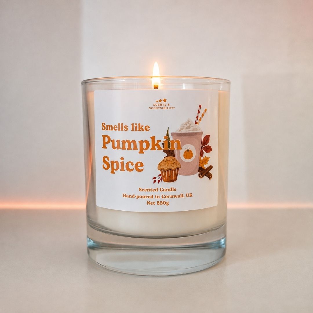 A lit scented candle in a clear glass jar with a white label that reads 'Smells like Pumpkin Spice' and shows autumn-themed illustrations. The candle is photographed on a neutral light beige surface against a matching background.