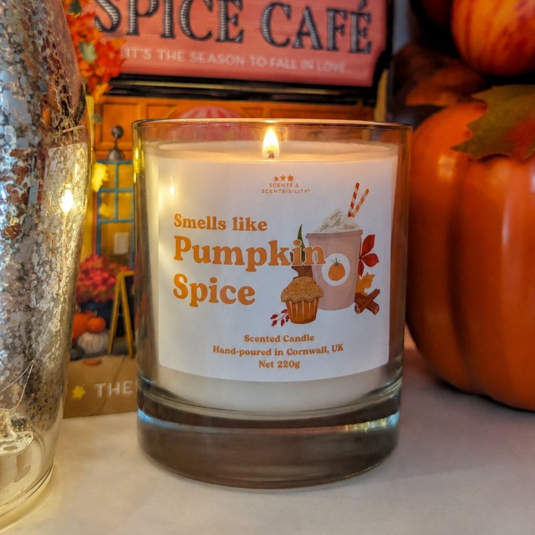 A lit candle in a glass jar labelled Smells Like Pumpkin Spice, surrounded by autumnal decor and books. 