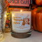 A lit candle in a glass jar labelled Smells Like Pumpkin Spice, surrounded by autumnal decor and books. 