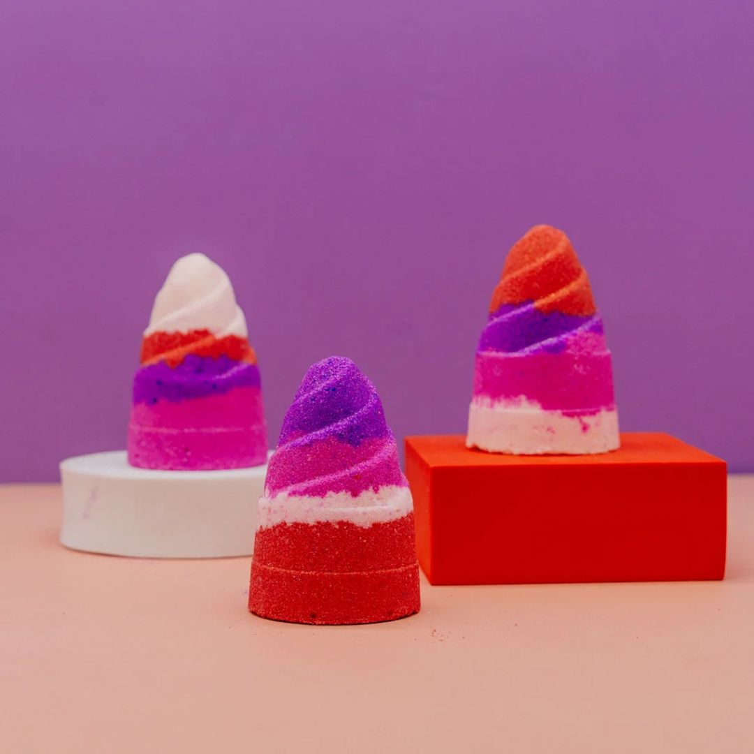 Three cone-shaped decorative bath bombs or sponges with layered candy-stripe colors, displayed on a pink surface against a purple background. One is on a white round pedestal, one in the center is red/purple/white, and one on the right is orange/purple/pink/white, all resting on red and white blocks.