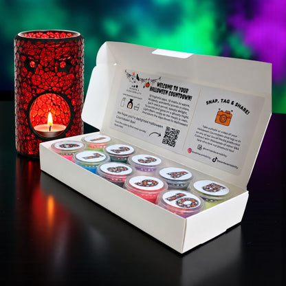 An open white box containing multiple small round pots of colorful wax melts, with a printed insert card inside the lid. A red crackle-pattern candle holder with a lit tea light candle sits beside the box on a dark reflective surface, against a colorful blurred background.