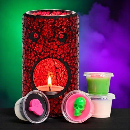 A Halloween-themed wax melt set featuring a red crackle-patterned wax melt burner with a lit tea light candle, alongside small plastic pots of green, pink, and white wax with skull and hand-shaped wax pieces in the lids, set against a green and purple smoky background.