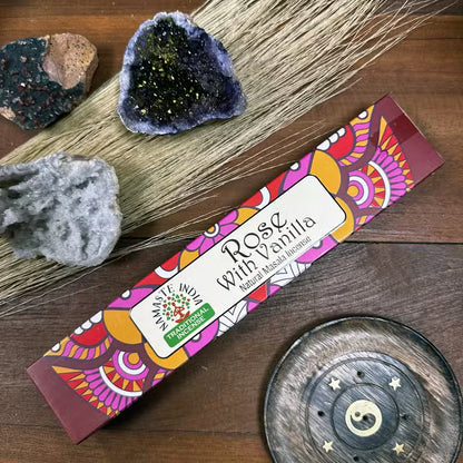 Colorful packaging of 'Rose with Vanilla' incense on a wooden surface with crystals and a decorative plate.