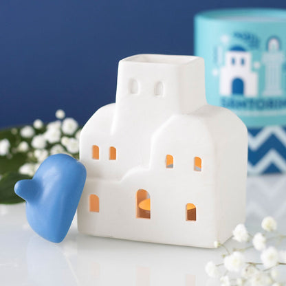 White ceramic Greek-inspired oil burner with a blue roof handle on a white surface with flowers.