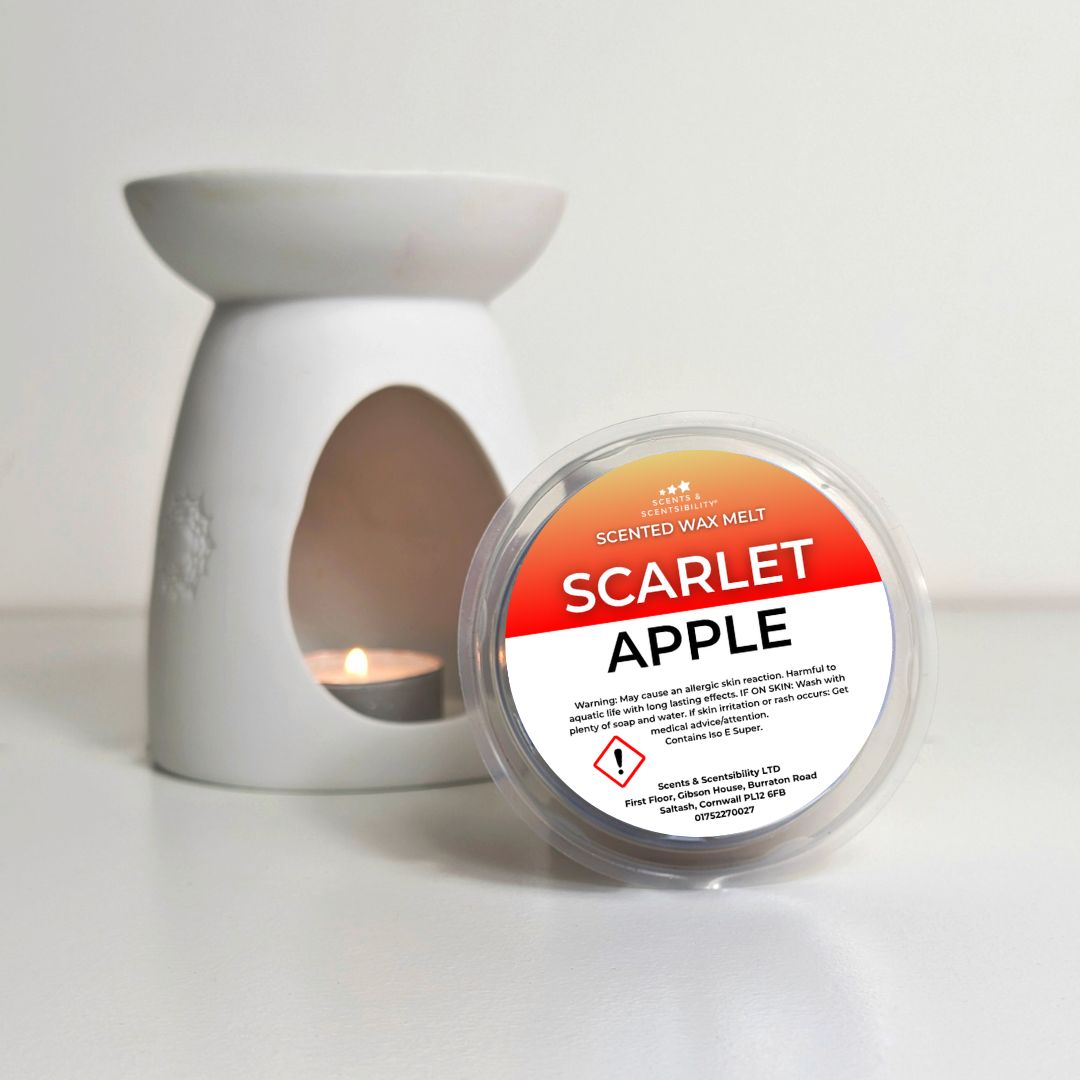 A round tub of scarlet apple scented wax melt from Scents of Calm, placed in front of a white ceramic candle warmer with a tea light candle lit inside, on a white surface.