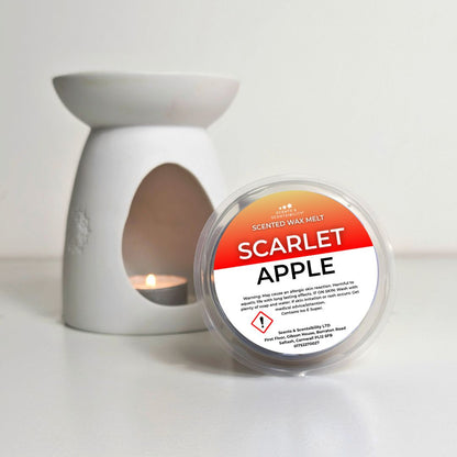 A round tub of scarlet apple scented wax melt from Scents of Calm, placed in front of a white ceramic candle warmer with a tea light candle lit inside, on a white surface.