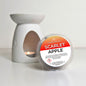 A round tub of scarlet apple scented wax melt from Scents of Calm, placed in front of a white ceramic candle warmer with a tea light candle lit inside, on a white surface.