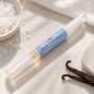 A clear cuticle pen with a brush on one side labeled 'SEA SALT AND VANILLA' with a light blue label, placed on a light stone surface with sea salt, a folded white towel, and vanilla beans nearby.