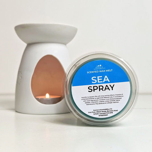 Scented wax melt container labeled 'Sea Spray' next to a lit candle in a white wax melt burner