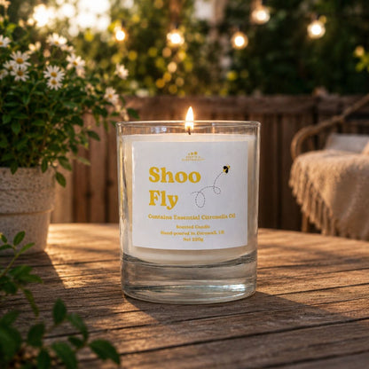 Candle labeled 'Shoo Fly' on a wooden table outdoors with plants and string lights in the background.