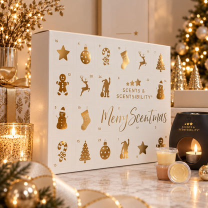 A white Scents & Scentsibility Christmas advent box with gold holiday icons and the text 'Merry Scentmas' sits on a marble surface surrounded by warm holiday decor, including lit candles, ornaments, and a blurred Christmas tree in the background.