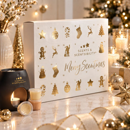 A white advent calendar box with gold holiday illustrations and the text 'Scented & Scentibility Merry Scentmas' sits on a tabletop with candles, ornaments, and warm festive decor in the background.