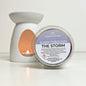 Scented wax melt container labeled 'Sleep Through The Storm' with a lit candle in the background.