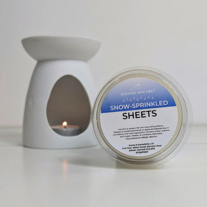 Scented wax melt container labeled 'Snow-Sprinkled Sheets' next to a white wax warmer with a lit candle.