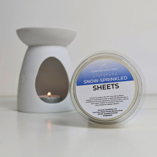 Scented wax melt container labeled 'Snow-Sprinkled Sheets' next to a white wax warmer with a lit candle.
