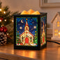 A square black wax melt warmer with stained-glass-style Christmas church panels, lit with warm yellow-orange wax and placed on a wooden table beside holiday decorations.