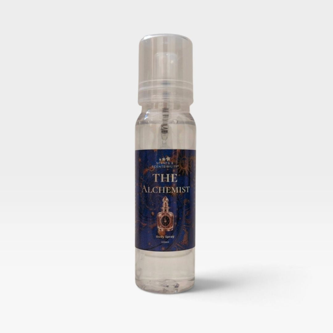 A clear plastic spray bottle of 'The Alchemist' body spray by Scents & Scentsibility, featuring a dark blue label with gold lettering and an illustration of an alchemy flask, set against a light gray background.