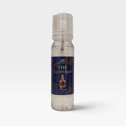 A clear plastic spray bottle of 'The Alchemist' body spray by Scents & Scentsibility, featuring a dark blue label with gold lettering and an illustration of an alchemy flask, set against a light gray background.