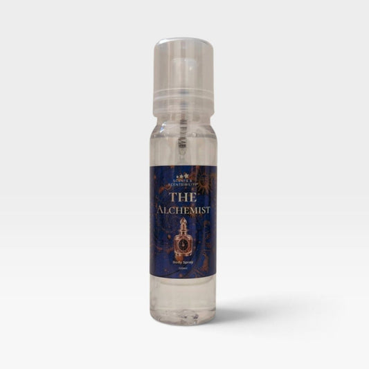 A clear plastic spray bottle of 'The Alchemist' body spray by Scents & Scentsibility, featuring a dark blue label with gold lettering and an illustration of an alchemy flask, set against a light gray background.