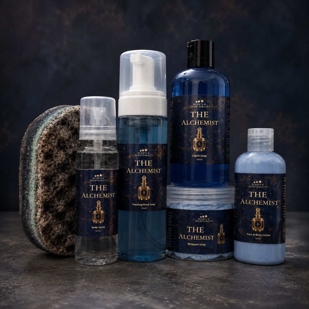 A set of six skincare products called 'The Alchemist' arranged on a dark textured surface against a dark background. The products include a body wash, foaming facial wash, body lotion, and a jar of whipped soap with one larger blue bottle and one smaller light blue bottle.