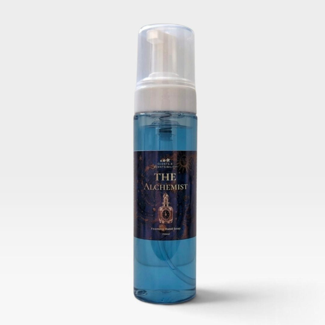 A bottle of The Alchemist Handmade Foaming Soap against a light gray background. The soap is a translucent blue liquid inside a clear bottle with a white pump dispenser and a dark navy label featuring gold text and illustrations.