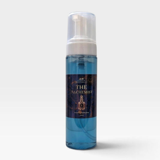 A bottle of The Alchemist Handmade Foaming Soap against a light gray background. The soap is a translucent blue liquid inside a clear bottle with a white pump dispenser and a dark navy label featuring gold text and illustrations.