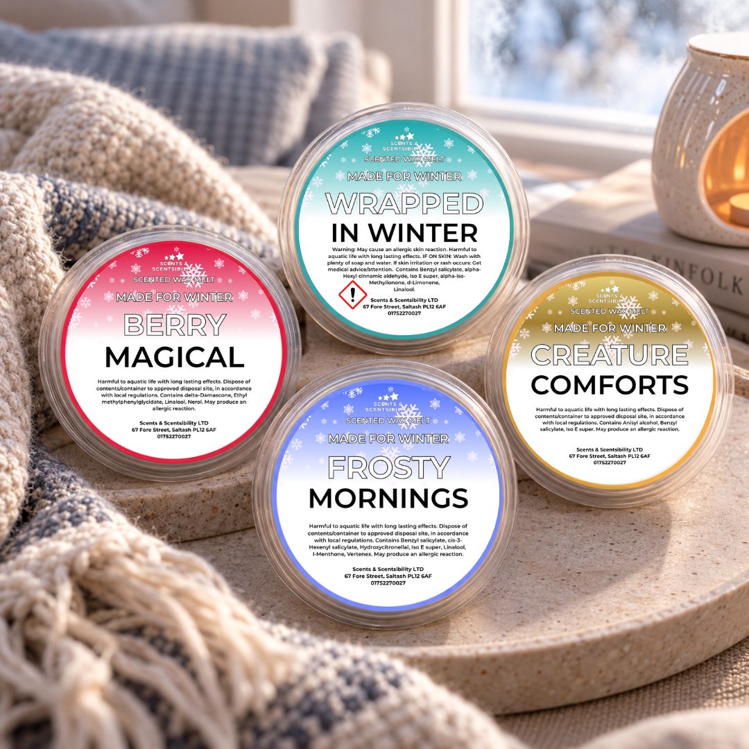 Four round wax melts in clear plastic tubs with colorful labels, arranged on a stone tray with a knitted blanket and a warm candle in the background. The labels read Berry Magical, Wrapped in Winter, Frosty Mornings, and Creature Comforts.
