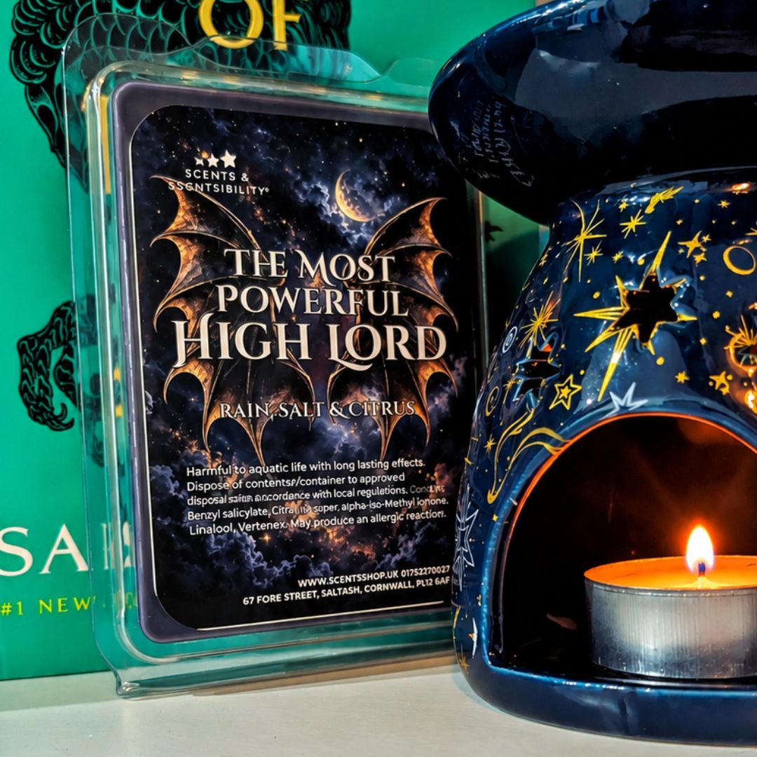 A wax melt package labeled 'The Most Powerful High Lord' by Scents & Scentsibility, shown next to a dark blue ceramic candle warmer with gold star and moon cutouts and a lit tealight candle inside.