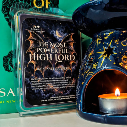 A wax melt package labeled 'The Most Powerful High Lord' by Scents & Scentsibility, shown next to a dark blue ceramic candle warmer with gold star and moon cutouts and a lit tealight candle inside.