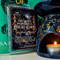 A wax melt package labeled 'The Most Powerful High Lord' by Scents & Scentsibility, shown next to a dark blue ceramic candle warmer with gold star and moon cutouts and a lit tealight candle inside.