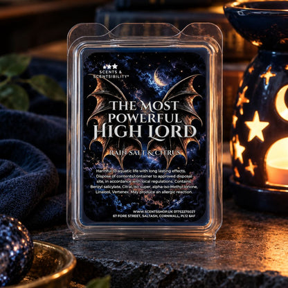 A wax melt package labeled 'The Most Powerful High Lord' by Scents & Scentsibility, shown in a clear clamshell container on a dark tabletop with warm ambient lighting and decorative lanterns in the background.