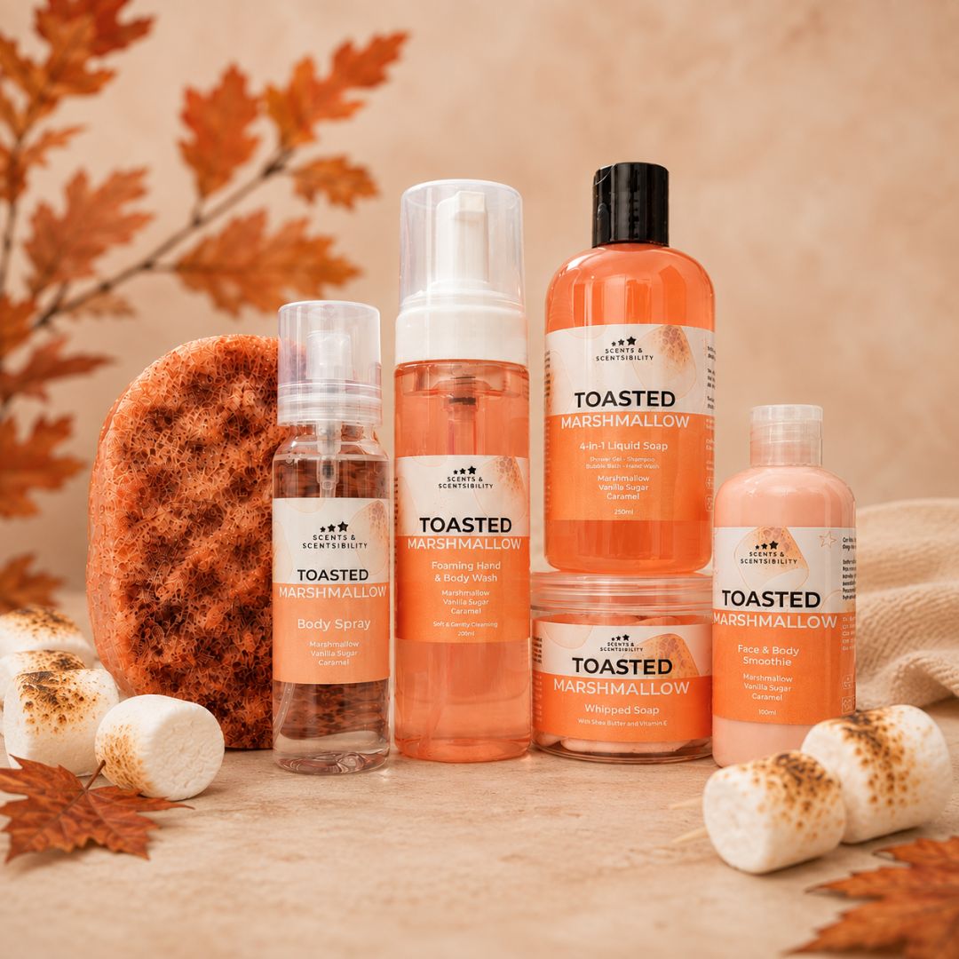 A set of five Toasted Marshmallow body care products arranged on a beige surface with autumn leaves, a marshmallow sponge, and toasted marshmallows. The products include body spray, body wash, hand wash, body butter, and foot and body wash in peachy orange and pinkish peach tones.