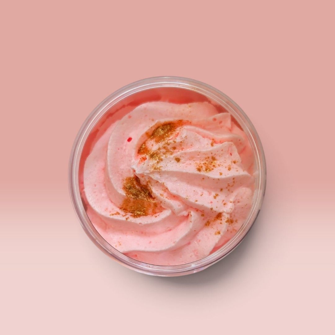 A round clear plastic tub filled with pink whipped soap, swirled on top of a soft pink background and lightly dusted with gold.