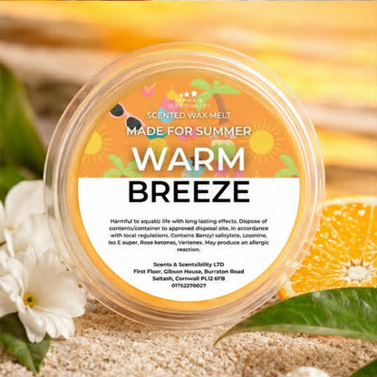 A round clear plastic tub of Scents & Scentsibility 'Warm Breeze' scented wax melt, featuring a white and orange label with illustrations of a beach umbrella, sunglasses, and sun, set against a tropical beach background with white flowers.