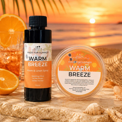 A bottle of room and linen spray and a round potof wax melts from Scents & Scentsibility labeled 'Warm Breeze', styled on a beach with orange slices, a drink, and a sunset background.