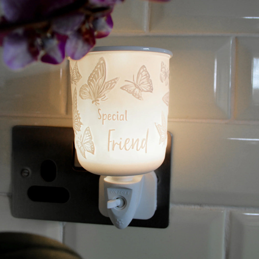 A butterfly-themed plug in wax melt warmer labeled 'Special Friend' glowing with a warm light, plugged into a black wall outlet against a white tiled wall.