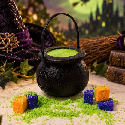 A black cauldron-shaped container filled with bright green bath bomb powder, styled on a tabletop with green sprinkles, orange and blue bath bomb cubes cubes, a witch hat, books, and a broom in a Halloween-themed setting.
