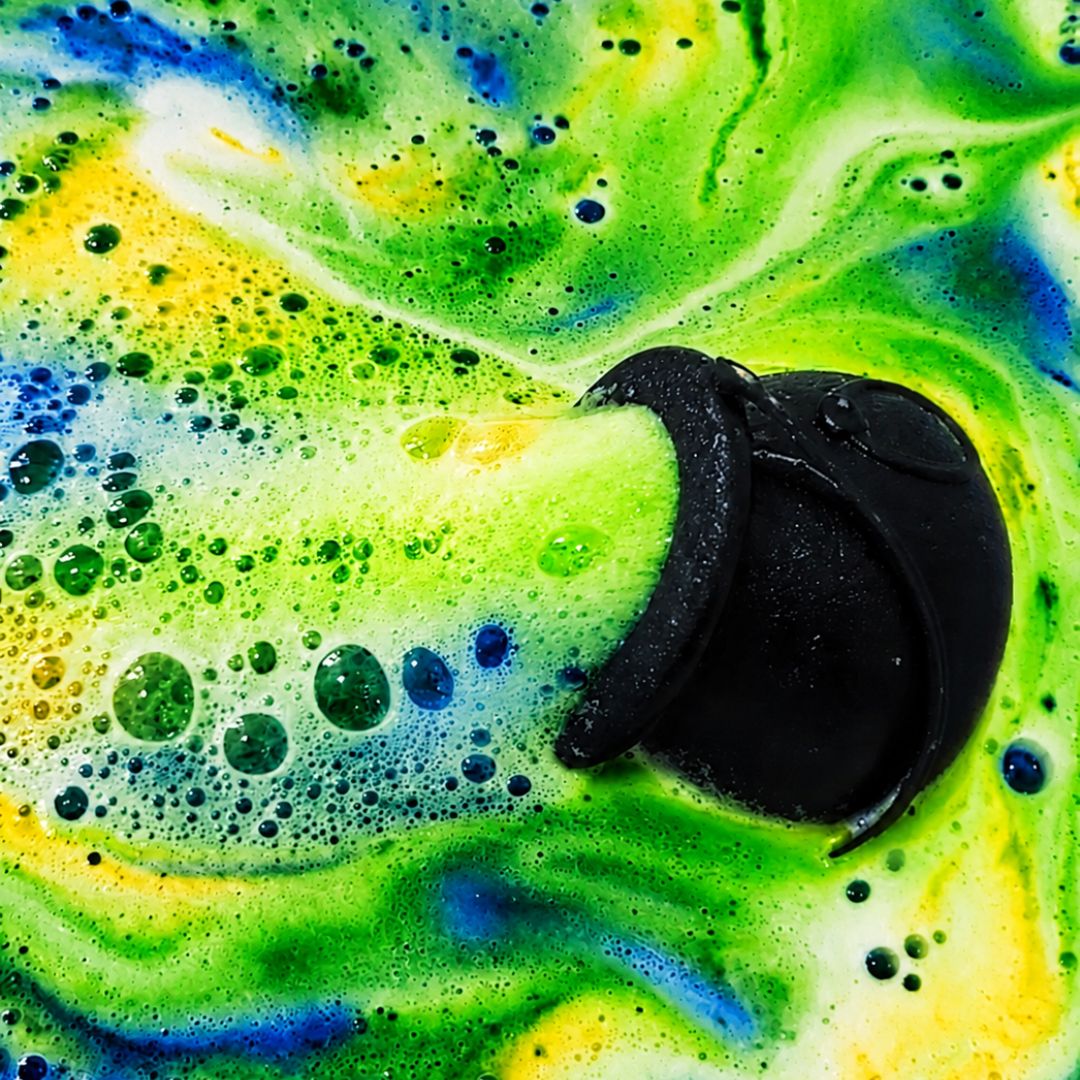A black cauldron bath bomb sits in a swirling, bubbly mixture of bright green, yellow, and blue foam.
